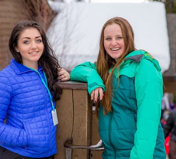 two teenage girls standing outside in winter coats leaning on a wooden post